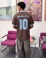 Camiseta de fútbol color marrón con cuello en V y detalles en celeste, inspirada en el Napoli F.C., con diseño de mangas cortas y sponsors impresos en el frente.