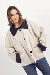 Chaqueta de corte holgado en color beige, con cuello y puños en contraste color azul marino. Presenta cierre frontal con botones oscuros y bolsillos laterales.