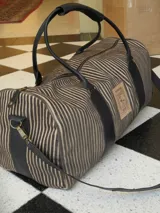 Bolso tipo duffel bag de viaje, confeccionado en tela con patrón de rayas verticales en tonos marrón y beige, con detalles y correas de cuero negro. Incluye asas de mano y correa de hombro desmontable.