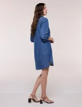 Vestido camisero blanco de rayón, con cuello con solapa, escote en V y pechera de abertura frontal con botones. Presenta un bolsillo delantero, bolsillos laterales aplicados y mangas largas con puño de largo regulable con presilla y botón. Cuenta con calce holgado y caída ligera, con ruedo irregular más largo en la parte trasera.