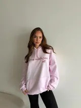 Hoodie canguro de felpa color rosado, con capucha ajustable con cordones y bolsillo delantero tipo kiko. Presenta un estampado frontal con la frase "All you need is love, self".