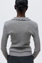 Buzo tipo polo de punto acanalado color gris, con cuello clásico, tapeta con tres botones nacarados y terminaciones con ribetes sobrehilados en puños y ruedo.