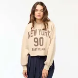 Buzo de cuello redondo en felpa color beige, con diseño estampado frontal con la inscripción 'NEW YORK 90 EAST VILLAGE' en color marrón. Presenta corte holgado y ruedo con terminación al corte.