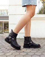 Bota de caña alta confeccionada en cuero negro, con suela track de goma. Presenta un diseño versátil con dos hebillas ajustables en la parte superior de la caña y una correa decorativa con tachas metálicas y argollas en la zona del tobillo.