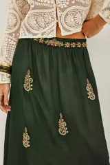 Vestido largo de satén color verde oscuro, con mangas tres cuartos terminadas en volados. Presenta un diseño con bordados dorados detallados en el pecho y cuello, además de motivos florales bordados dispersos a lo largo de la falda. Incluye un cinturón fino con aplicaciones brillantes.