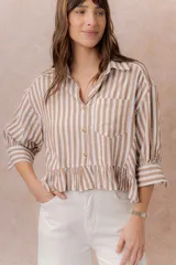 Camisa de rayas verticales blancas y marrones, de calce relajado y largo corto con volado en el ruedo. Presenta cuello camisero clásico, mangas tres cuartos con puños amplios, abotonadura frontal y un bolsillo aplicado en el frente.