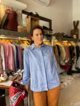 Camisa de algodón liviano color celeste, con manga larga, cuello camisero, bolsillo frontal con parche y etiqueta de marca, y pespuntes en contraste. Cuenta con cierre frontal oculto y botones de madera y coco.