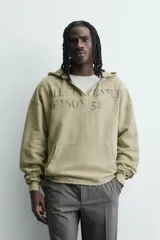 Sudadera de corte relajado color verde oliva con efecto lavado. Presenta capucha con abertura frontal, bolsillo tipo canguro, mangas largas con puños de rib y bajo con acabado irregular. Incluye estampado frontal con texto.