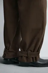 Pantalón de algodón color marrón oscuro, estilo balloon fit, con talle alto, cierre frontal y trabillas ajustables en los bajos. Presenta costuras verticales marcadas en el frente de cada pierna.