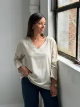 Blusa de lino color beige con escote en V, mangas largas con sutiles frunces en los hombros y costuras decorativas visibles en el cuello y el ruedo.