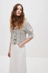 Blusa color gris claro en tejido de mezcla de viscosa y algodón, con escote en V pronunciado, volados frontales, alforzas, inserciones de encaje y detalles bordados. Cuenta con mangas abullonadas al codo con puños elásticos de volado y volado ancho en el bajo.