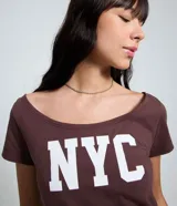 Remera de algodón color marrón oscuro con escote amplio tipo barco u hombro a hombro y manga corta. Presenta el estampado en mayúsculas "NYC" en color blanco en el frente.