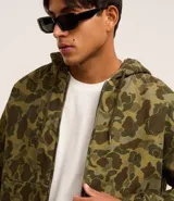 Campera con capucha y cierre frontal, confeccionada en sarga con estampado camuflado. Tiene manga larga con sisa desplazada, puños ajustados, bolsillo canguro y bajo con elástico.