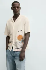 Camisa de manga corta y calce relajado en color beige, con cuello bowling y cierre frontal de botones. Presenta un diseño bordado en la parte inferior con motivos de paisaje tropical que incluyen palmeras, sol y aves.
