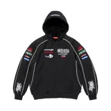 Campera Supreme de estilo racing en color negro, con capucha forrada en corderito negro, cierre frontal completo y bolsillos tipo canguro. Presenta diversos parches bordados con temática automovilística en el pecho y las mangas, además de líneas blancas decorativas.