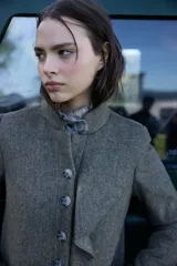 Chaqueta de lana en tono verde oliva con textura jaspeada, cuello mao y detalle de volado frontal. Presenta un corte entallado, bolsillos delanteros con solapa y cierre frontal con botones.