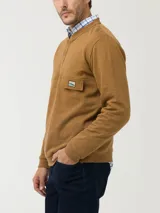 Buzo de algodón orgánico color tostado, con manga larga, cuello redondo y cierre parcial con broches a presión. Presenta un bolsillo aplicado en el pecho con solapa y un parche con diseño de paisaje de la marca RKF.