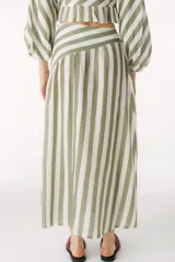 Falda larga color beige de lino, con corte amplio y vuelo, que presenta un canesú diagonal en la parte superior que genera volumen fruncido en la falda.