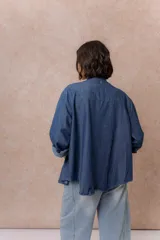 Camisa de denim azul con bordados florales en tonos amarillos y celestes sobre los hombros y el pecho. Presenta cuello clásico, cierre frontal con botones y mangas largas con puños.