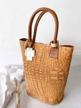 Bolso tipo tote de cuero color marrón con textura tipo cocodrilo, doble asa corta de cuero marrón y detalle de borlas decorativas.