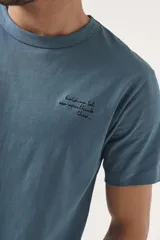 Remera de manga corta color azul grisáceo, con cuello redondo y un pequeño texto bordado en negro en el pecho.