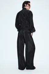 Pantalón de tiro alto color verde militar, confeccionado en tejido satinado con acabado brillante. Presenta cintura elástica fruncida, bolsillos laterales ocultos en las costuras y perneras de corte ancho.