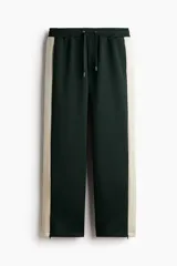 Pantalón deportivo de corte relajado en tejido efecto neopreno, color verde oscuro con franjas laterales en contraste color crema. Presenta cintura elástica con cordón ajustable, bolsillos laterales y traseros, y cierre oculto en el bajo.