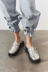 Mocasines tipo mule de cuero metalizado plateado, con suela track negra y detalle de tira con hebilla en el empeine.