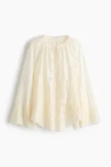 Blusa de lyocell color marfil con diseño de broderie tono sobre tono, cuello redondo fruncido, escote en V con cordón para atar y mangas largas raglán con ribetes festoneados.