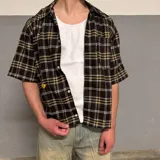 Camisa de manga corta con corte boxy fit, diseño a cuadros en tonos marrones, amarillos y azules, con cierre frontal de botones y bolsillo en el pecho.