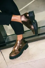 Botines de cuero metalizado en tono oscuro con acabado brillante. Presentan un diseño de corte bajo con una hebilla lateral decorativa y suela de goma negra con plataforma dentada.