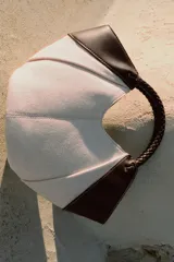 Bolso mini shopper de lona en color crudo con detalles en cuero sintético marrón en los laterales y una manija corta trenzada de mano. Cuenta con costuras contrastantes y cierre magnético.