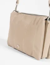 Bandolera de tamaño compacto en color beige, confeccionada en cuero vegano. Presenta un diseño con pespuntes verticales visibles, cierre superior y correa larga ajustable y extraíble. Incluye un pequeño aplique metálico decorativo en la parte inferior frontal.