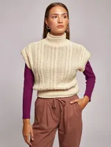 Chaleco tejido de punto color beige, con cuello alto y hombreras marcadas. Presenta un diseño de trenzas verticales en el frente y un ajuste elástico en la cintura.