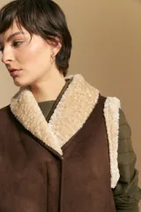 Chaleco negro de gamuza sintética con interior y bordes de corderito sintético. Presenta un diseño sin mangas con cuello solapa y cierre frontal.