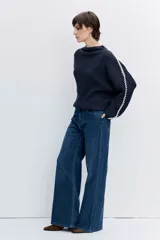 Jersey de punto color marrón chocolate, de cuello alto y corte holgado. Presenta un diseño acanalado y un detalle de costura decorativa en color blanco que recorre la parte superior de las mangas.