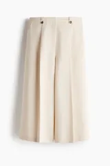 Pantalón sastrero cropped de tiro alto, color beige, con piernas amplias, pliegues frontales, bolsillos al bies y trabillas en la cintura con detalles de botones.