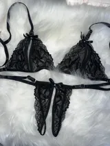 Conjunto de lencería de dos piezas confeccionado en encaje negro, compuesto por un soutiene triangular con aberturas frontales y una bombacha tipo colaless abierta con detalles de moños y apliques de strass.