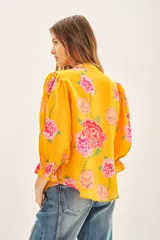 Camisa de viscosa con estampado floral en tonos amarillos y rosas. Presenta escote redondo con cierre frontal de botones, mangas al codo con terminación elástica y detalle de volado.