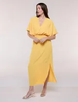 Vestido largo amarillo de corte fluido con escote en V, mangas cortas amplias y apliques de tachas en el escote y puños. Posee cintura elastizada fruncida y falda larga con tajos laterales en el ruedo.