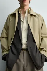 Trench de corte relajado confeccionado en tejido técnico italiano, color beige. Presenta cuello camisero, manga larga, bolsillos de vivo en la cadera y cierre frontal de botonadura simple con botones oscuros.