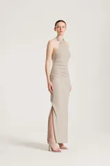 Vestido largo de fiesta color dorado con acabado brillante, escote halter y detalle de pedrería en el cuello. Presenta fruncidos en el lateral y una abertura en la falda.