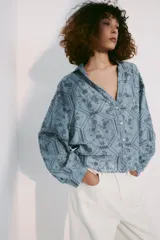 Camisa azul con cuello de solapa, escote en pico y mangas largas con puños abotonados. Presenta un diseño con bordados perforados y patrones geométricos y florales. Cierre frontal con botones.