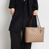 Bolso tote grande de Guess, color marrón con estampado logomarca en patrón repetitivo. Presenta asas de hombro en color marrón oscuro y herrajes dorados. Incluye un dije colgante con el logo de la marca.