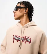 Buzo canguro beige de algodón fleece con capucha y cordón ajustable. Presenta un bordado frontal con el texto "Forged by Fire" en tipografía estilo gótico con bordes rojos y negros.