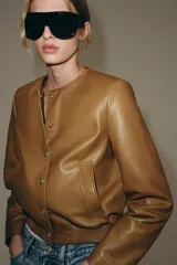 Campera bomber de cuero 100% color tostado de la ZARA WOMAN COLLECTION, con cuello redondo, manga larga, bolsillos delanteros ribeteados y cierre frontal con botones metálicos en relieve. Presenta elástico fruncido en el dobladillo inferior.