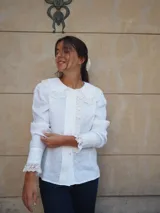 Camisa de lino color beige, con cuello bobo y detalles de encaje bordado en el cuello, pechera y puños. Cuenta con mangas largas levemente abuchonadas y cierre frontal con botones nacarados.
