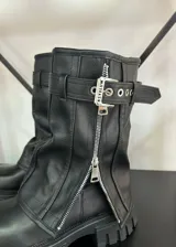 Botas de caña media color negro, con suela track dentada, cierre frontal con cremallera metálica y tira con hebilla ajustable en la parte superior.