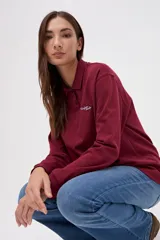 Remera tipo polo de manga larga en color beige, confeccionada en algodón pesado con textura suave. Presenta cuello clásico, cierre frontal con botones y un pequeño bordado en el pecho. Posee puños en las mangas y un corte relajado.