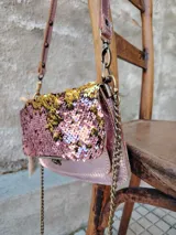 Cartera bandolera con solapa, color rosa con lentejuelas doradas y plateadas. Tiene correa de cuero sintético rosa y cadena dorada.